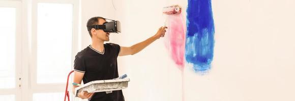 Young man with dark hair is painting the wall in white in his apartment. In his hands he holds a paint roller and paint brush. Can not decide what to choose. photo