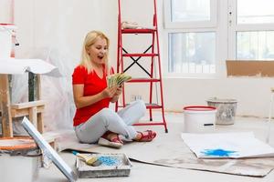 Woman holds bundle of dollars cash money, sits on floor with instruments for renovation apartment isolated on white background. Wallpaper accessories for gluing painting tools. Concept of repair home photo