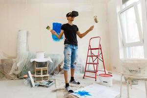 joven hombre con oscuro pelo es pintura el pared en blanco en su departamento. en su manos él sostiene un pintar rodillo y pintar cepillar. lata no decidir qué a elegir. foto