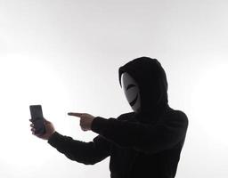 hacker anónimo y mascarilla con smartphone en la mano. hombre con camisa de capucha negra sosteniendo y usando un teléfono móvil con fondo blanco. representan el concepto de piratería informática o robo de datos personales foto