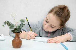 pequeño niña sentado a el mesa con concentración y diligentemente escribe en un computadora portátil, espalda a colegio foto