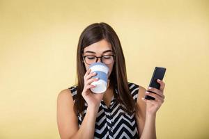 Portrait of cheerful youth holding hot beverage and drinking coffee while holding smart mobile phone isolated over peach background. Smiling girl in with take away coffee cup and cell phone . photo