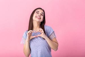 Portrait of a smiling young beautiful woman showing heart gesture with two hands and looking at camera isolated over pink background photo