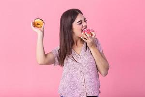 Beauty model girl eating colorful donuts. Funny joyful styled woman choosing sweets on pink background. Diet, dieting concept. Junk food, Slimming, weight loss photo