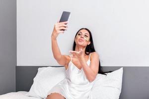 Young happy woman with Gold Cosmetics mask, Eye Patches using mobile phone for video call , gesturing kisses to friends. Beautiful girl making facetime video calling with smartphone lying on the bed. photo