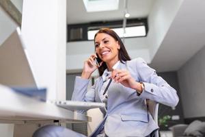 Professional businesswoman working at her office via laptop, young female manager using portable computer device while sitting at modern loft and talking on her mobile phone, work process concept photo