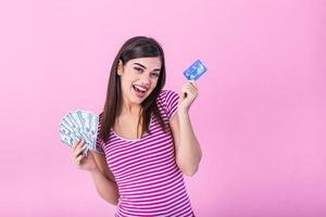 retrato de un niña en un raya camiseta en pie en un rosado antecedentes. modelo mira a el cámara demostración banco tarjeta y un haz de dolares con soñador Mira foto