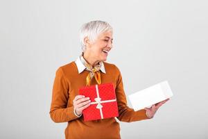 Portrait of a happy smiling mature woman opening a Christmas gift box isolated over gray background. Excited senior casual woman holding present box photo