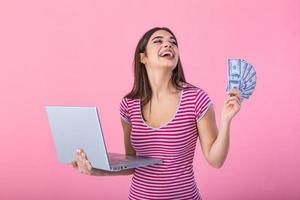 Portrait of excited satisfied girl holding money banknotes with laptop computer isolated over pink background. Portrait of a cheerful young woman holding money banknotes and laptop computer in hands photo