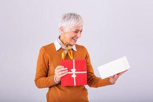 Portrait of a happy smiling mature woman opening a Christmas gift box isolated over gray background. Excited senior casual woman holding present box photo