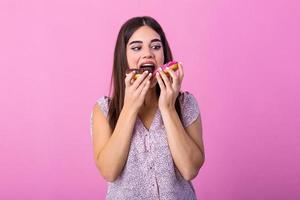 Stylish girl with long hair positively poses, holding fresh pink and chocolate donuts with powder ready to enjoy sweets..Close up portrait of a satisfied pretty girl eating donuts photo