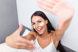 hermosa joven mujer con oro productos cosméticos mascarilla, ojo parches sentado en cama y muestra gesto foto marco con dedos. retrato de sonriente dama posando en brillante hogar . concepto felicidad y amor.