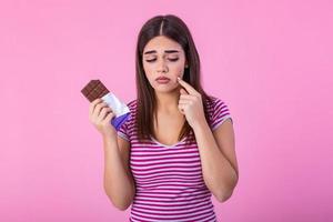 Teenage girl with acne problem holding chocolate bar against pink background. Young beautiful Woman Acne Problem Face with Chocolate bar UnHappy Eating,His Stressful face. photo