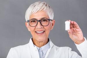 Portrait of a confident mature female doctor dressed in uniform with stethoscope pointing finger at bottle with pills isolated over dark background photo