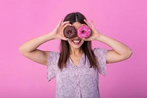 Close up portrait of a cute girl posing with donuts at her face isolated over violet background. Beautiful woman with donuts on her eyes photo