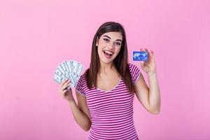 retrato de un niña en un raya camiseta en pie en un rosado antecedentes. modelo mira a el cámara demostración banco tarjeta y un haz de dolares con soñador Mira foto