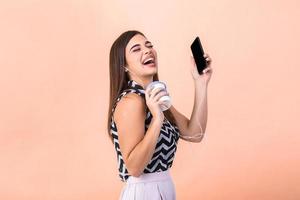 Portrait of excited girl enjoy coffee to-go in her best mood. Young attractive female holding smartphone and cup of coffee to go and laughing photo