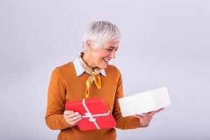 Portrait of a happy smiling mature woman opening a gift box isolated over gray background. Excited senior casual woman holding present box photo