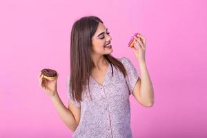 Beauty model girl eating colorful donuts. Funny joyful styled woman choosing sweets on pink background. Diet, dieting concept. Junk food, Slimming, weight loss photo