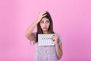 Portrait of a beautiful young girl holding her menstruation calendar and covering hear face isolated over pink background. Period calendar photo