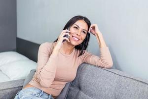 sonriente hermosa mujer hablando en el teléfono a hogar, contento joven niña sostiene Teléfono móvil haciendo respondiendo llamar, atractivo niña teniendo agradable conversacion chateando por móvil con amigo foto