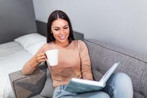 Happy young beautiful woman relaxing at home in cozy winter or autumn weekend with book and cup of hot tea or coffee, sitting in sofa and reading a book photo
