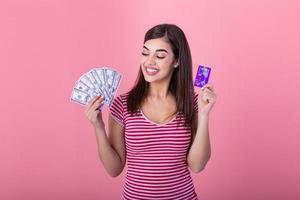 Photo of caucasian brunette woman 20s smiling while holding credit card and fan of dollar cash isolated over pink background