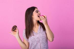 Beauty model girl eating colorful donuts. Funny joyful styled woman choosing sweets on pink background. Diet, dieting concept. Junk food, Slimming, weight loss photo