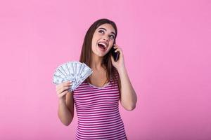 imagen de una joven mujer bonita sorprendida gritando sorprendida posando aislada sobre fondo de pared rosa usando un teléfono móvil con dinero. foto