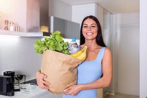 retrato de hermosa joven mujer mirando a cámara mientras participación compras bolso con Fresco vegetales y frutas comestibles en el cocina a hogar. foto