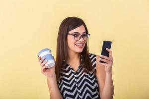 Indoor picture of young good-looking European woman isolated on peach background holding coffe to go and phone in hand and smiling while browsing or reading message photo