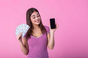 Excited attractive woman holding smartphone and money, cashback. Image of excited young lady isolated over pink background. Showing display of mobile phone holding money. photo