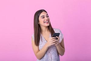 Adorable girl with mobile phone in hand expressing positive emotions. Attractive woman in pink shirt holding smartphone and texting on studio pink background photo