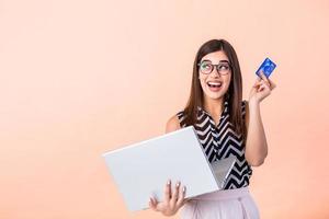 Portrait of a joyful young woman using laptop computer, showing credit card isolated over background. Young woman holding credit card and using laptop computer. Online shopping concept. photo