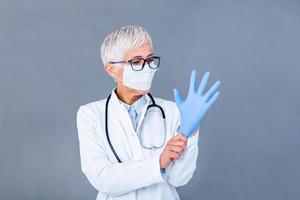 Mature female Doctor putting on protective gloves and medical protective mask, isolated on background. Doctor putting on sterile gloves photo