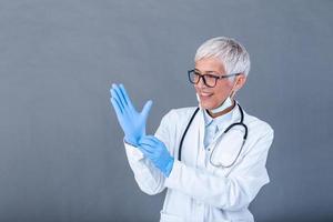 Mature female Doctor putting on protective gloves and medical protective mask, isolated on background. Doctor putting on sterile gloves photo