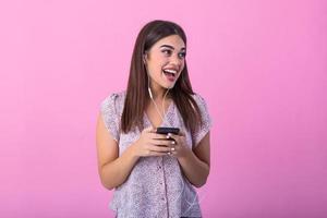 Excited beautiful girl hold mobile phone with headphones listening to music on blank empty screen isolated on pink studio background . Emotions, lifestyle concept. Copy space.