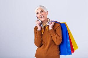Photo of happy amazing mature woman standing isolated over white wall background. Looking aside holding shopping bags while chatting on her mobile phone.
