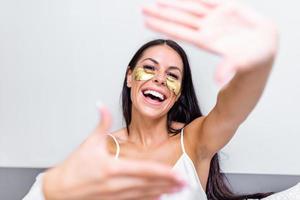 Beautiful young woman with Gold Cosmetics mask, Eye Patches. sitting on bed and shows gesture photo frame with fingers. Portrait of smiling lady posing in bright home . Concept happiness and love.