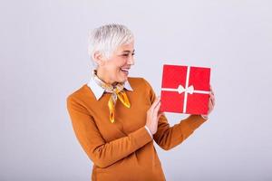 Portrait of cheerful positive glad charming aged woman with hairstyle having gift box in red package with white bow enjoying holiday festive mood isolated on grey background photo