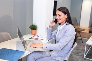 Professional businesswoman working at her office via laptop, young female manager using portable computer device while sitting at modern loft and talking on her mobile phone, work process concept photo