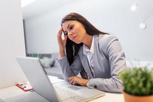 Young frustrated woman working at office desk in front of laptop suffering from chronic daily headaches, treatment online, appointing to a medical consultation, electromagnetic radiation, sick pay
