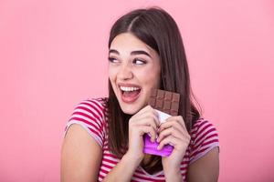 Happy young beautiful lady eating chocolate and smiling. Girl tasting sweet chocolate. Young woman with natural make up having fun and eating chocolate isolated on pink background photo