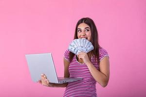 retrato de un emocionado satisfecho niña participación dinero billetes con ordenador portátil computadora aislado terminado rosado antecedentes. retrato de un alegre joven mujer participación dinero billetes y ordenador portátil en manos foto