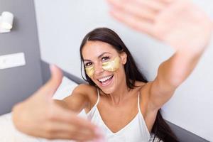 hermosa joven mujer con oro productos cosméticos mascarilla, ojo parches sentado en cama y muestra gesto foto marco con dedos. retrato de sonriente dama posando en brillante hogar . concepto felicidad y amor.