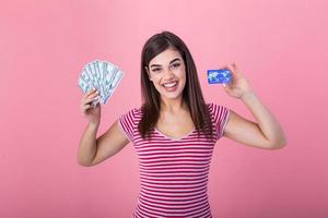 Portrait of a girl in a stripe t-shirt standing on a pink background. Model looks at the camera showing bank card and a bundle of dollars with dreamy look photo