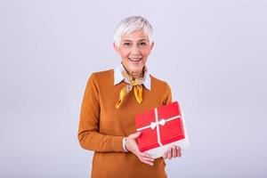 retrato de alegre positivo alegre encantador Envejecido mujer con peinado teniendo regalo caja en rojo paquete con blanco arco disfrutando fiesta festivo estado animico aislado en gris antecedentes foto