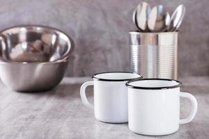 Two white empty metal mugs, spoons in a can and metal plates on a gray background photo