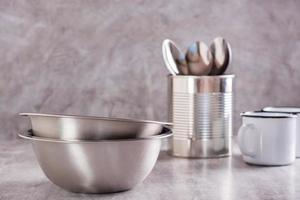 Two empty metal bowls, spoons in a can and metal enamel mugs on a gray background photo