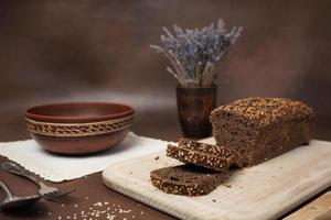 Black Bread with cereals Sliced on a Wooden cutting Board against a brown background. Next to the bread is a Clay Plate, fork, spoon and white cotton tablecloth photo